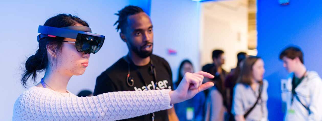 A woman wearing special goggles interacts with an augmented reality interface while a man looks on with interest.